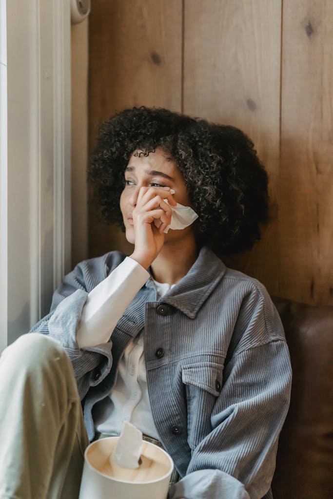 Unhappy young ethnic woman with Afro hair in casual clothes wiping tears with tissue while sitting near window and crying