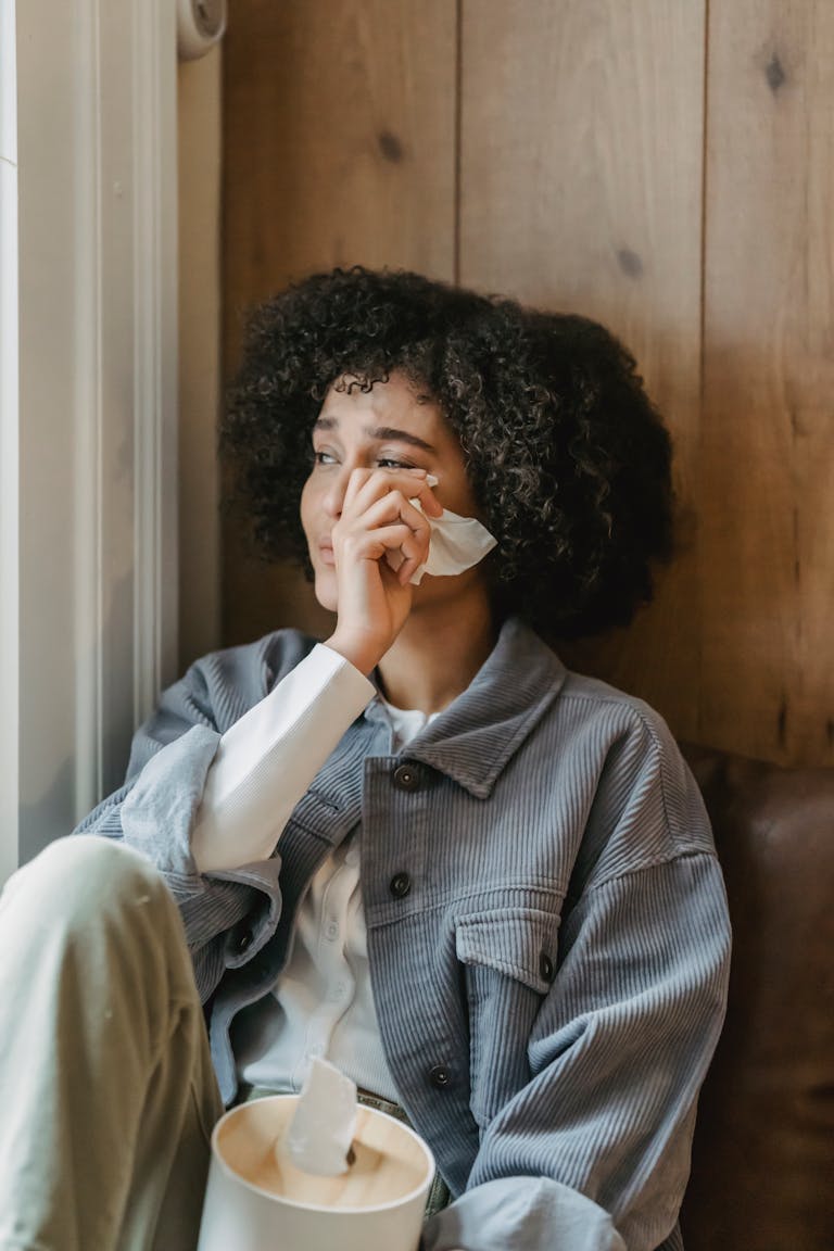 Unhappy young ethnic woman with Afro hair in casual clothes wiping tears with tissue while sitting near window and crying